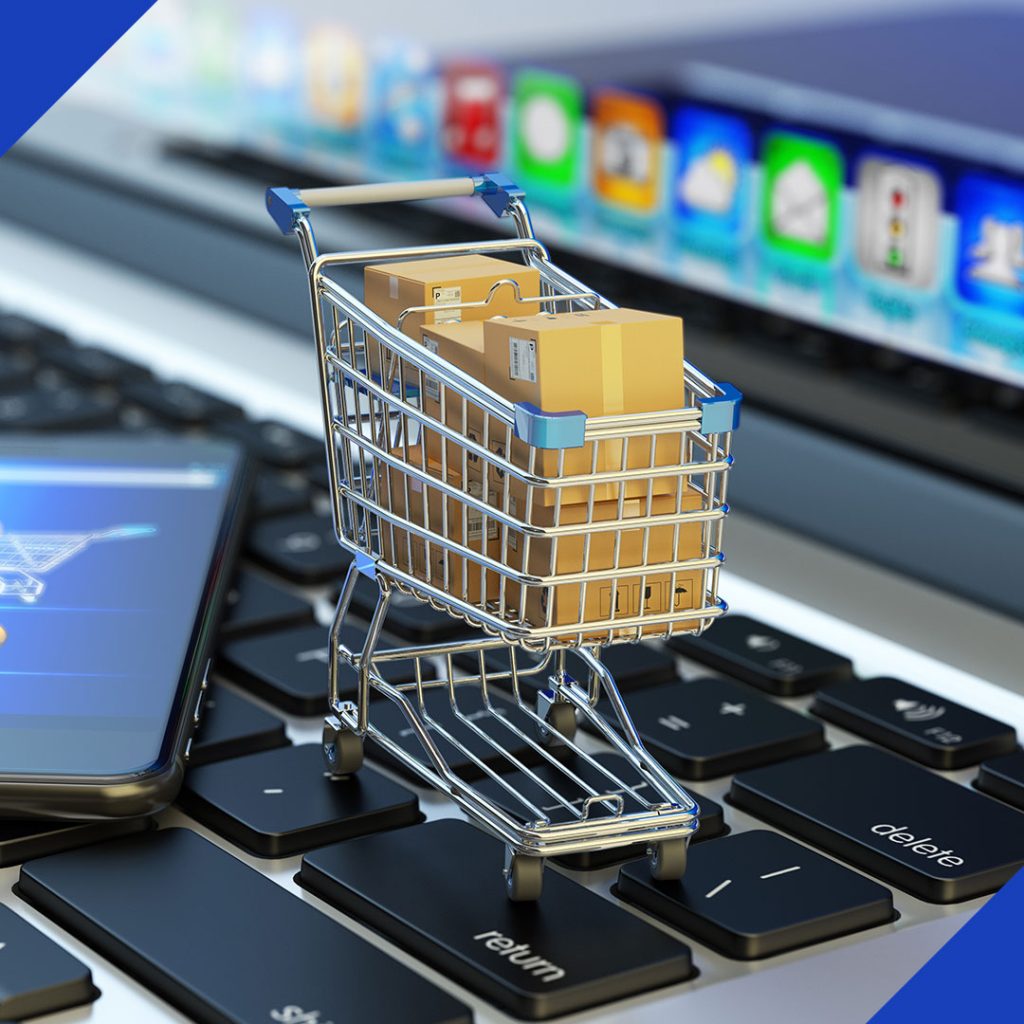 A miniature shopping cart with cardboard boxes on a laptop keyboard next to a smartphone, symbolizing the e-commerce platform services from JR Digital Media.